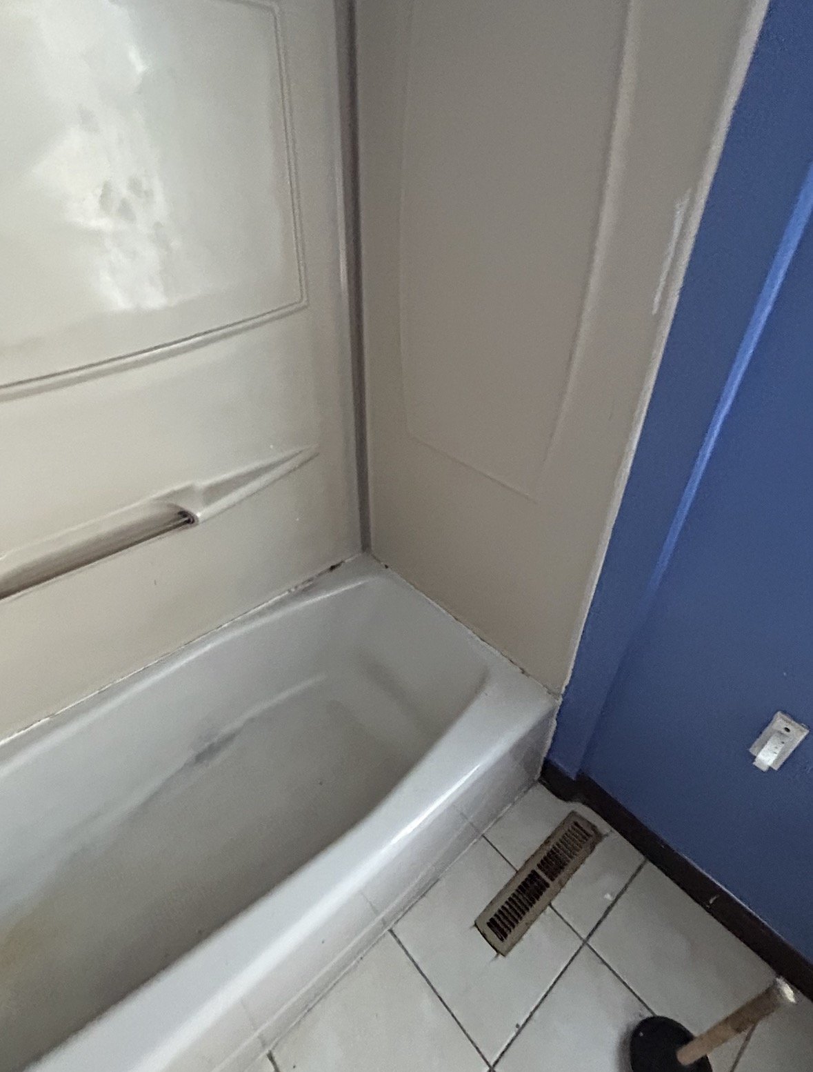 Close-up of the corner of a shower stall, with a drain on the white tiled floor nearby, blue painted wall, and a white wall with an electrical outlet.