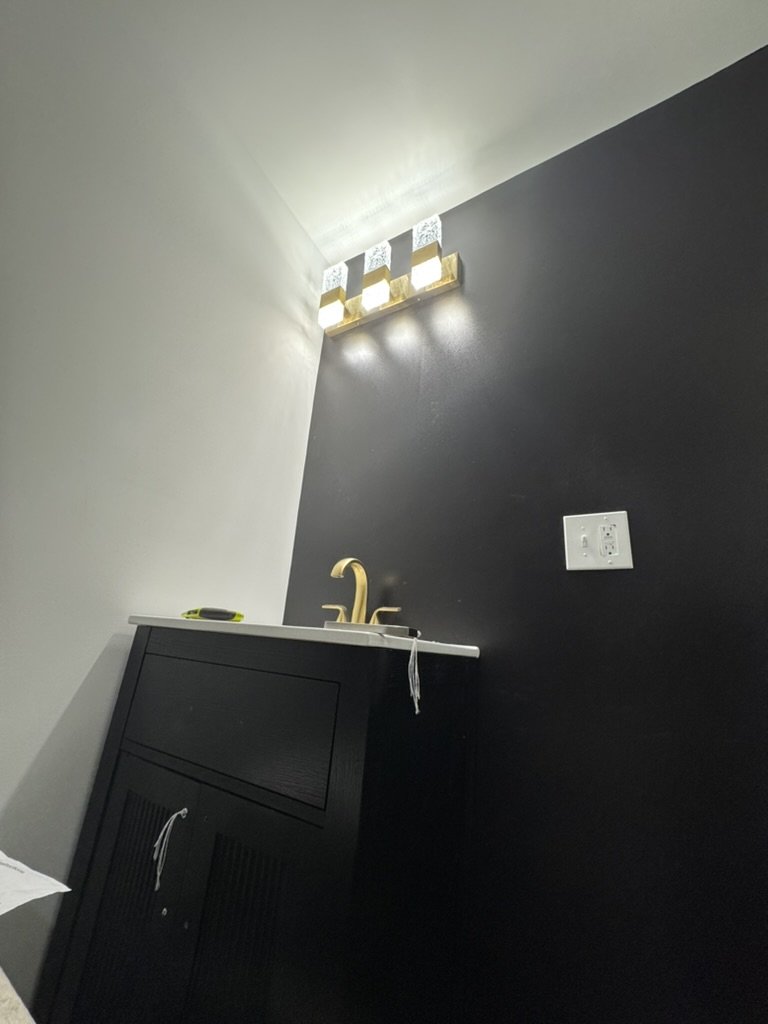 A black bathroom vanity with a gold faucet and a white countertop, mounted on a black wall. Above the vanity, there is a modern light fixture with multiple glass blocks emitting light. An electrical outlet is on the black wall next to the vanity.