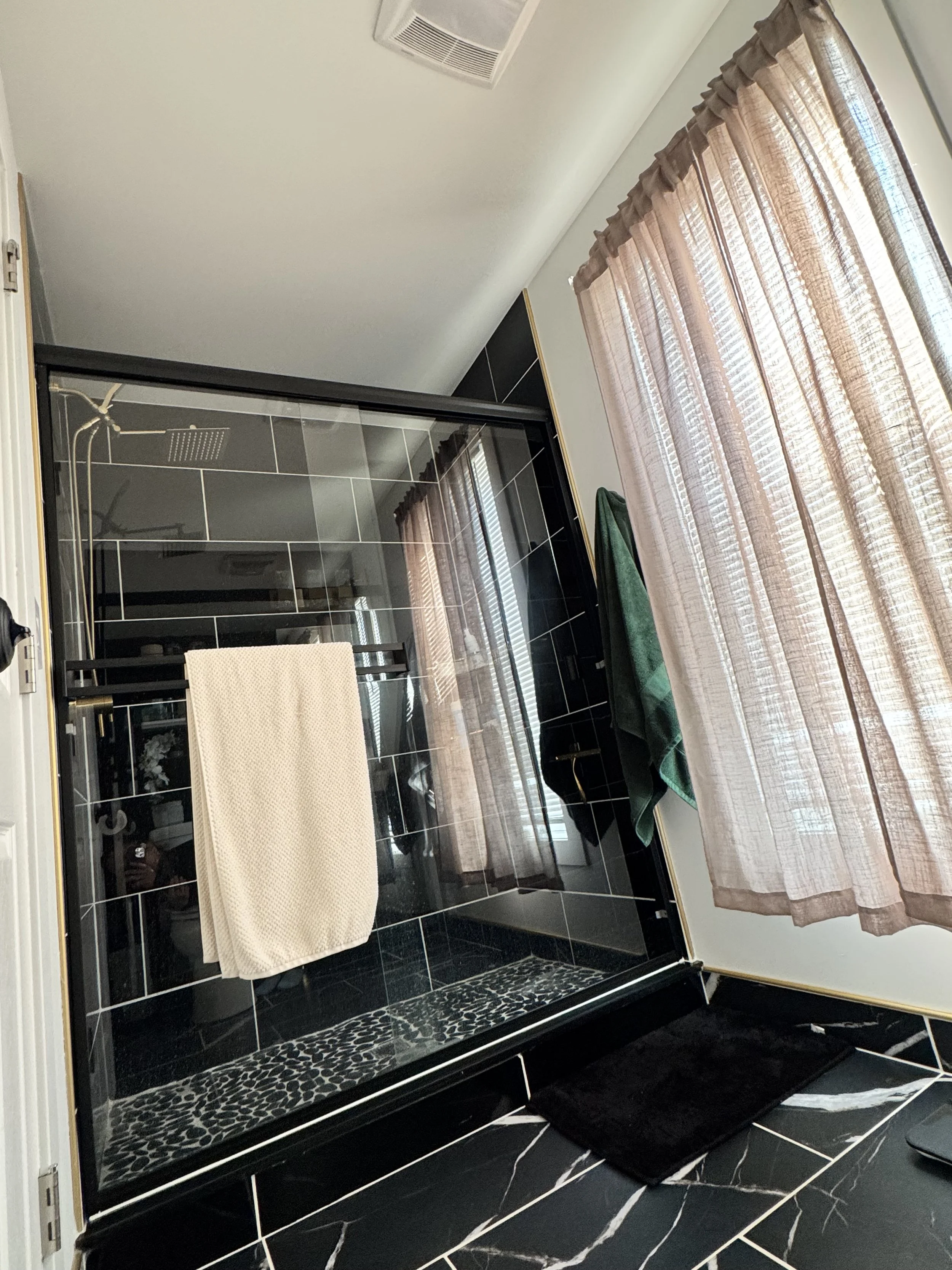 A bathroom with a glass shower enclosure, black tiled floor with white veining, beige curtains on a window, and two towels hanging—one white inside the shower and one green outside.