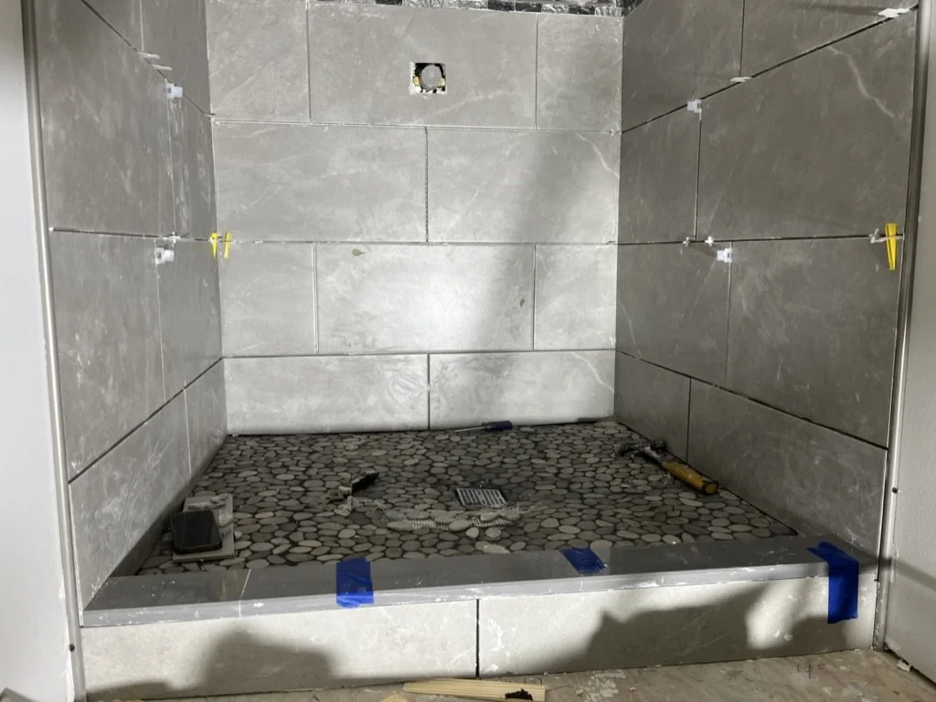 A shower stall under construction with beige tiles on the walls, small pebble-like tiles on the floor, and construction tools and materials inside.