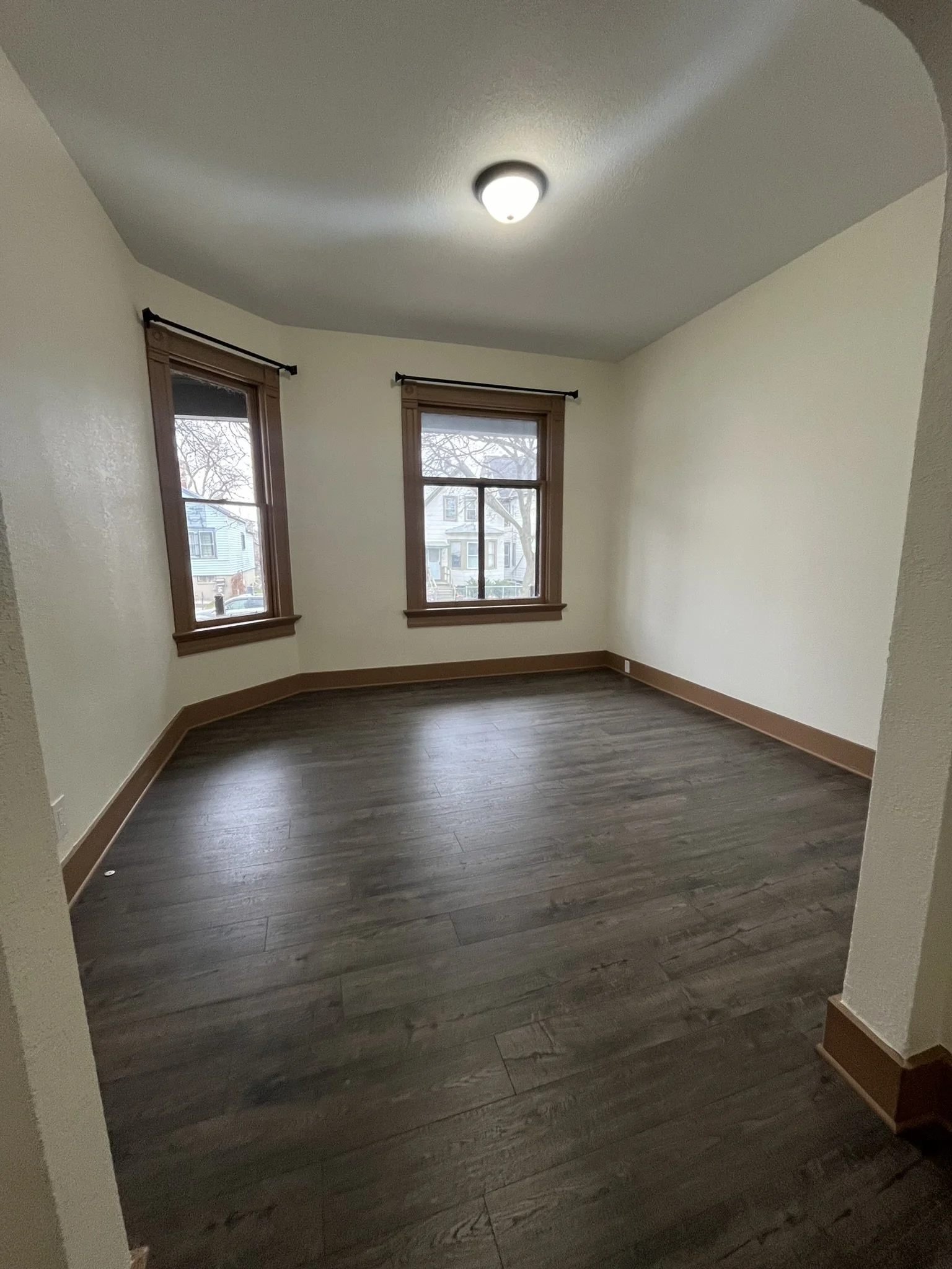 Empty room with three windows and dark wood flooring, cream walls, wooden trim, and an overhead light.