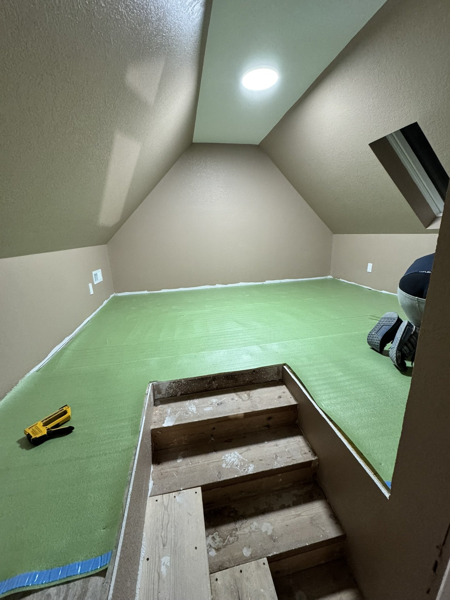 Attic room under renovation with drywall installed on the sloped ceiling and walls, a bright ceiling light, green flooring, and construction tools like a screwdriver and kneepads.