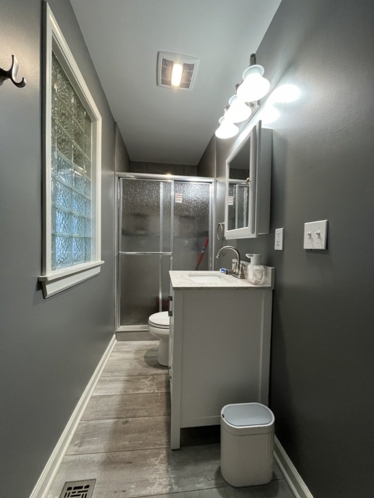 A small bathroom with dark gray walls, a window with glass blocks, a metal-framed shower with a frosted glass door, a white toilet, a white vanity with a sink, a mirror cabinet above the sink, bright light fixtures, and a trash bin on the floor.