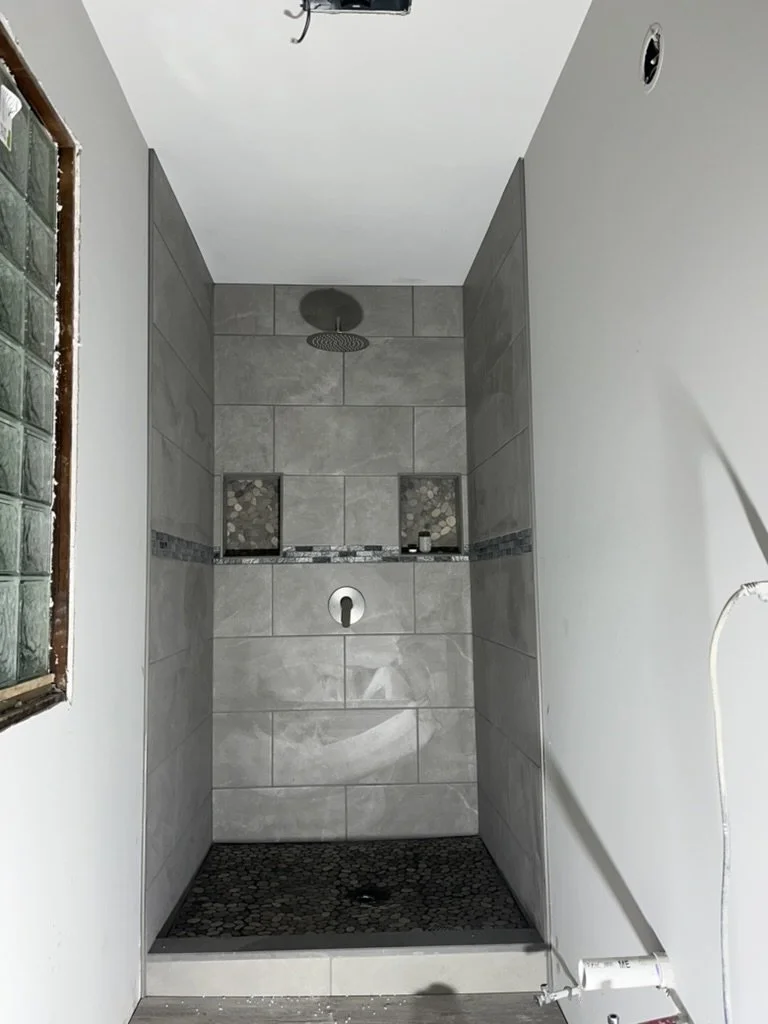 A walk-in shower with gray tiled walls, two built-in niches for toiletries, a round showerhead, and a black pebble floor. The shower is unfinished, without a door or curtain.