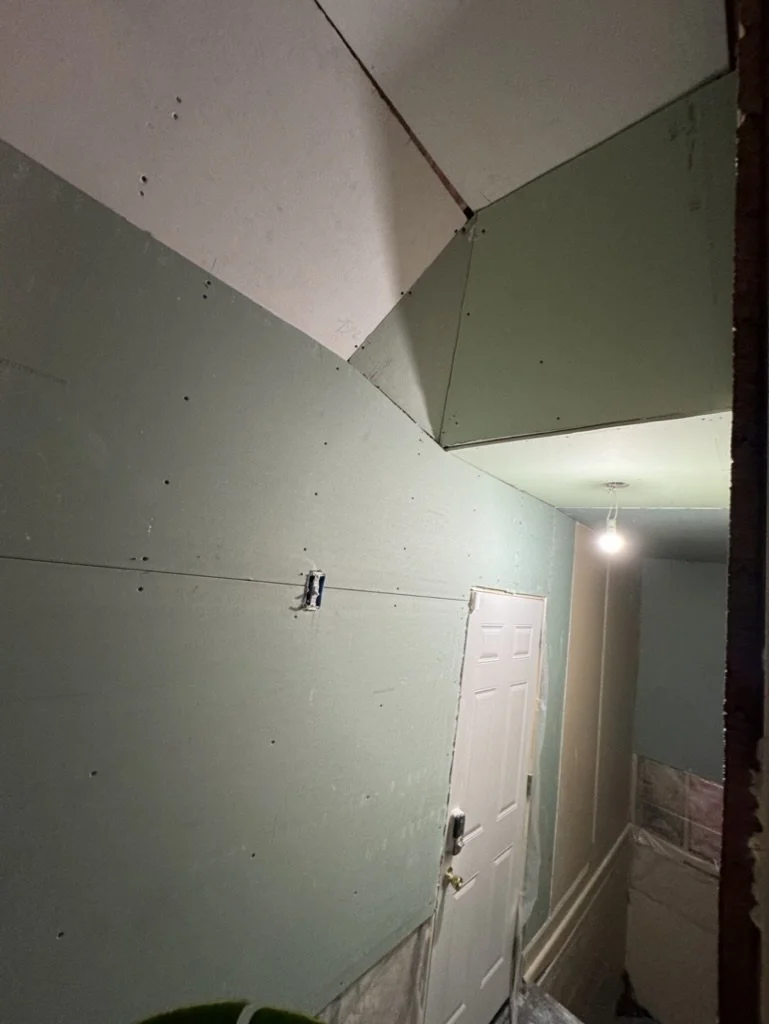 A narrow hallway under construction with unfinished drywall, a closed white door, and an unfinished ceiling with exposed drywall panels and a hanging light bulb.
