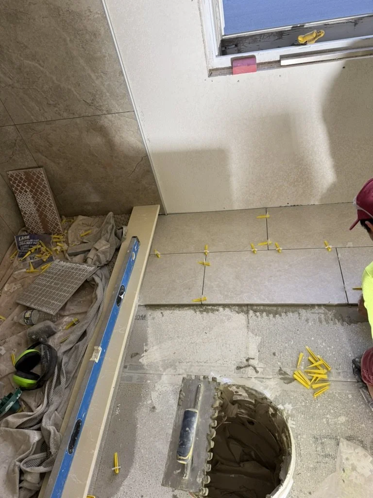 Construction workers installing tiles in a room, with tools, spacers, and a bucket of mortar visible.