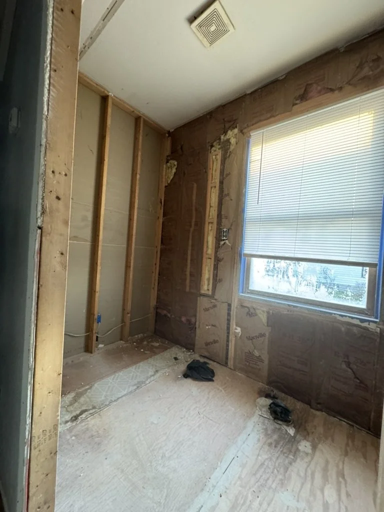 A room under renovation with exposed wall studs and insulation, a window with blinds, and a vent on the ceiling.