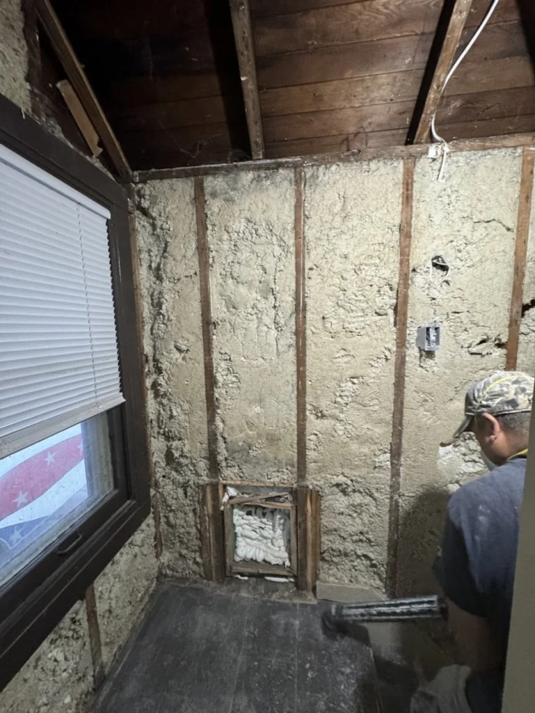 Person working on a wall exposed with wall insulation, removing drywall, with window to the left and a ceiling with visible wooden beams at the top.