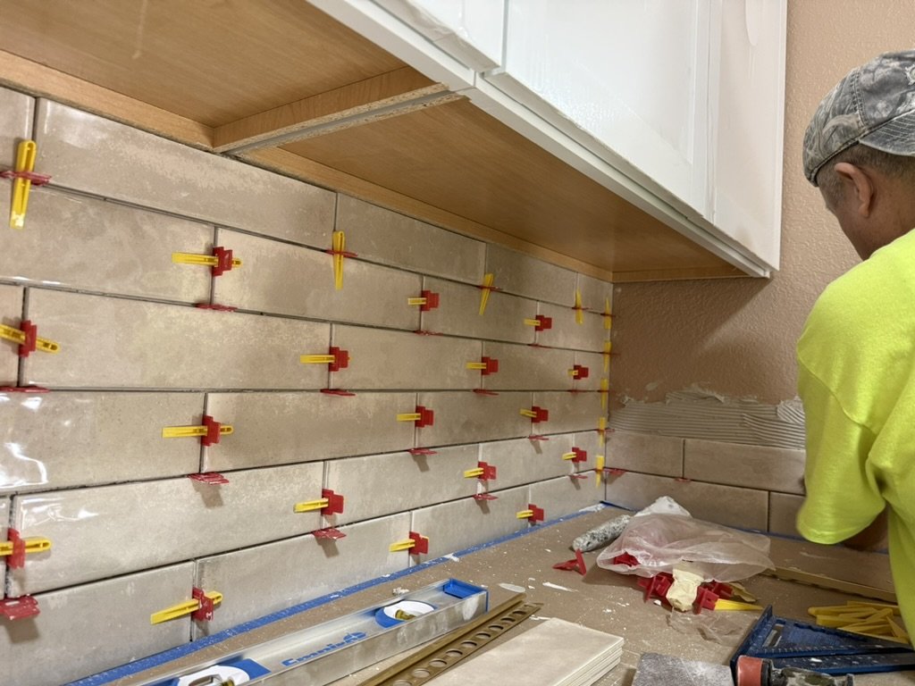 Person installing ceramic tiles on kitchen wall, using clamps to hold tiles in place during installation.