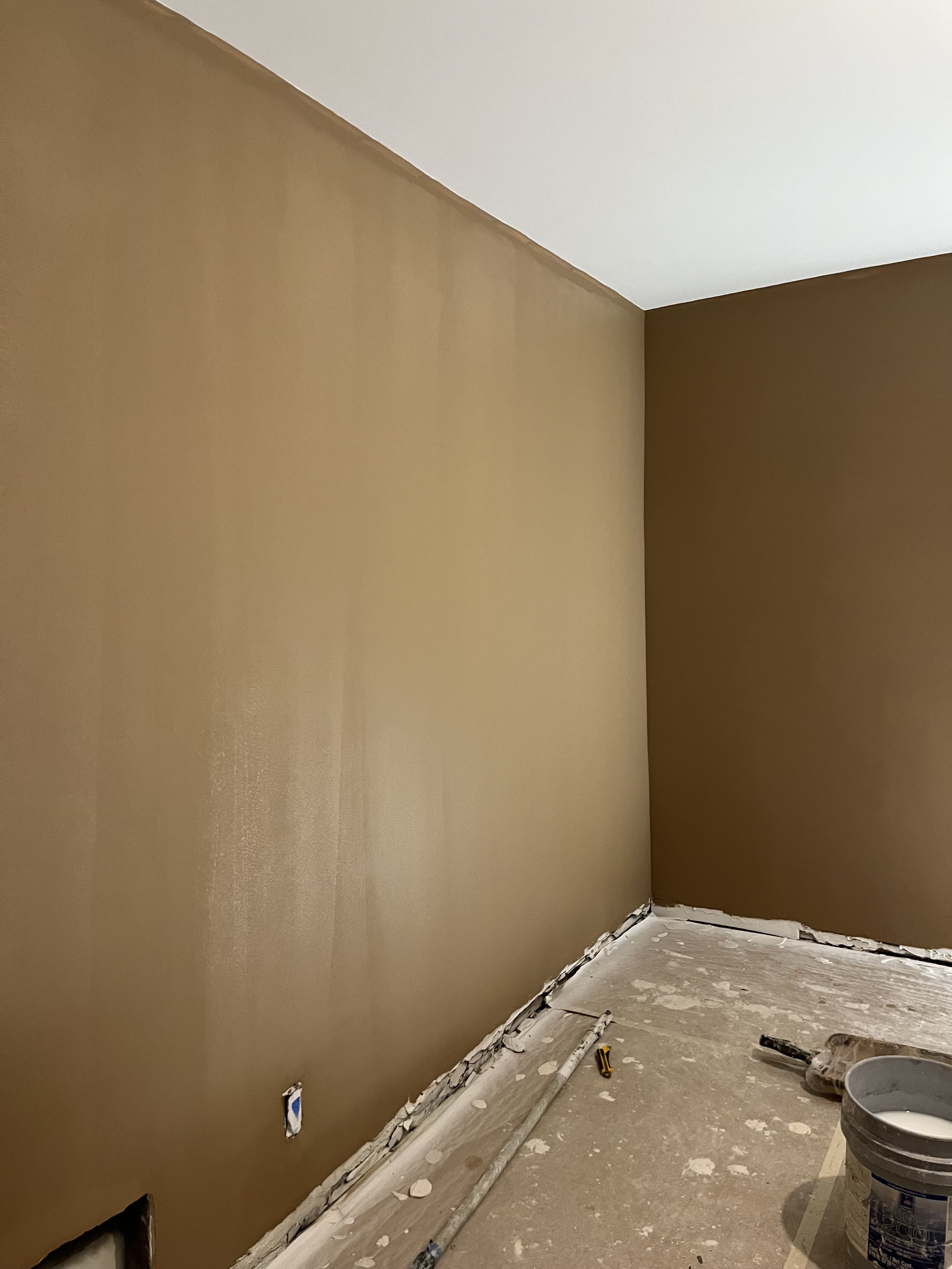 A room with walls painted in two shades of brown, with flooring under renovation. There is a bucket, a paint roller, and a level on the floor, and part of the wall has been freshly painted with some painter's tape around an electrical outlet.
