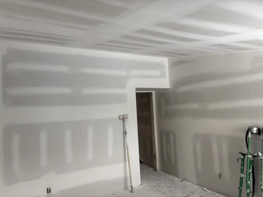 Drywall installation in a room, with visible joint compound patches and a doorway, construction tools including a drywall knife and a scaffold present.