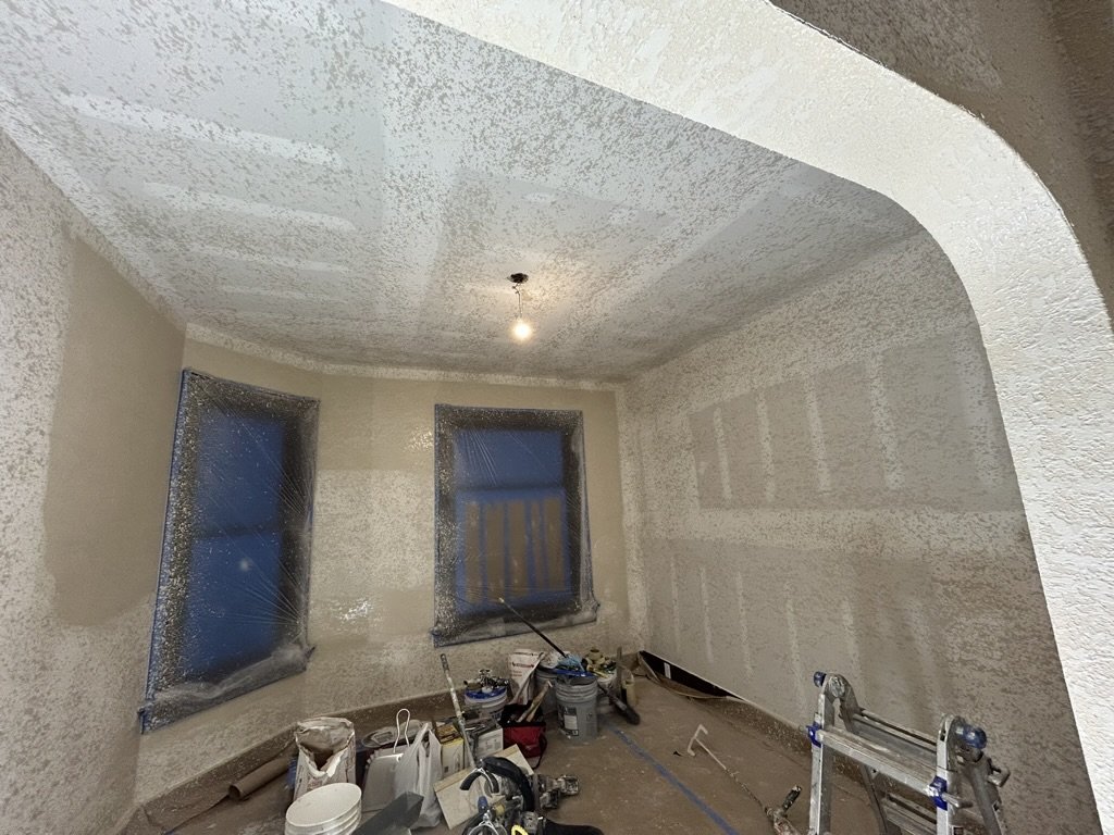 A room under construction with textured beige walls and ceiling, two covered windows, construction tools, and materials on the floor.