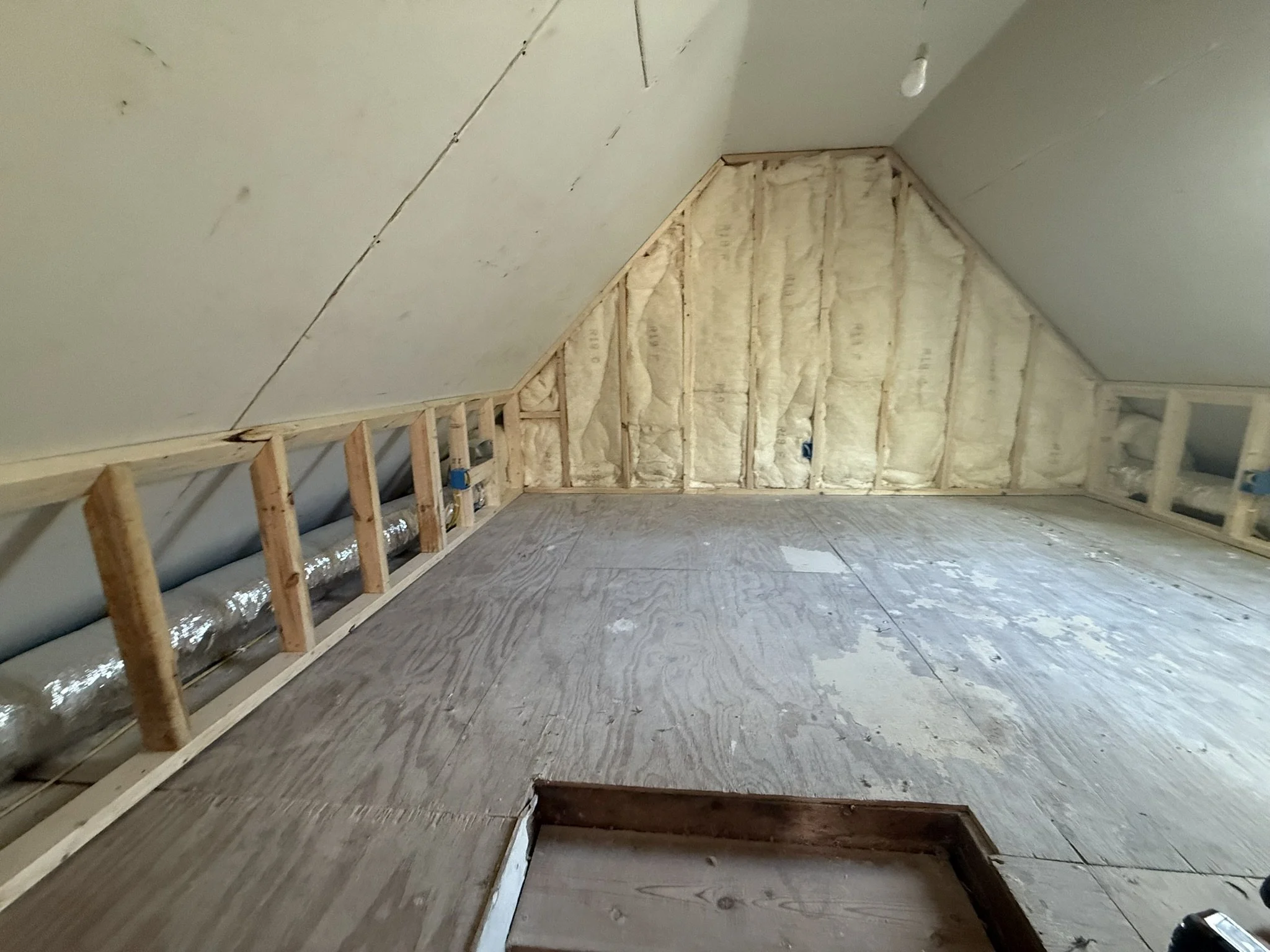 An attic space under construction with exposed insulation and wooden framing, unfinished walls, and a plywood floor.