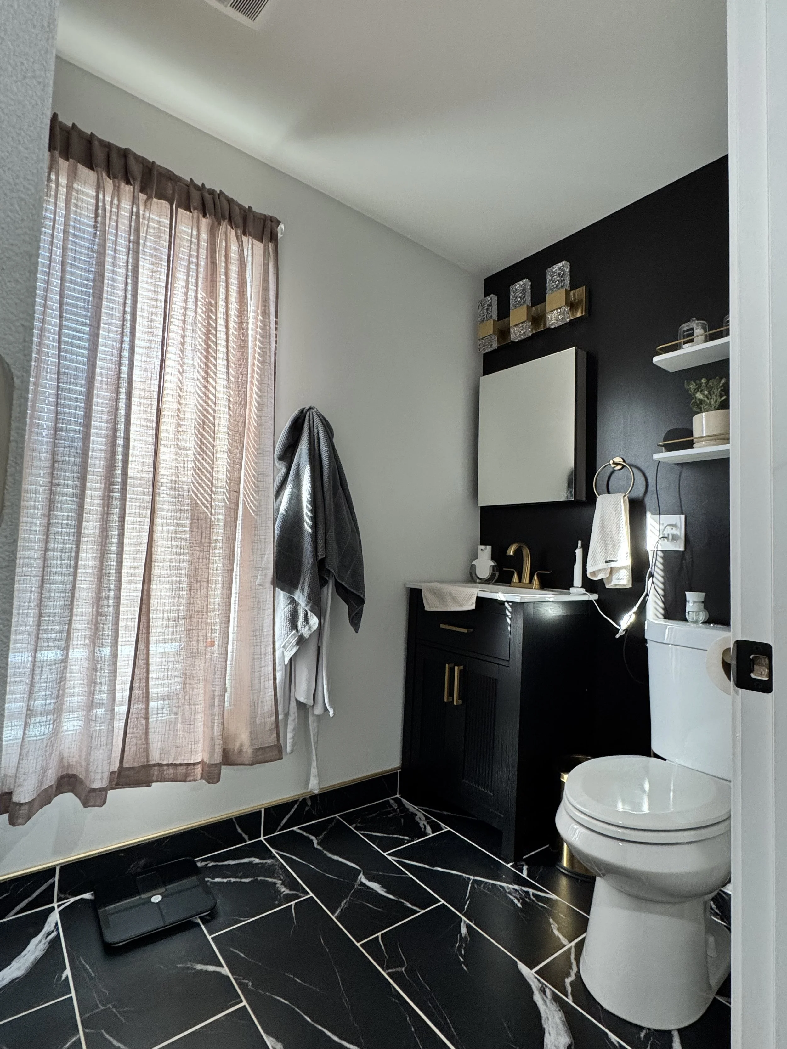 A bathroom with black marble tile flooring, a window with pink curtains, a black vanity with gold handles, a white toilet, and decorative wall shelves with plants and glass containers.