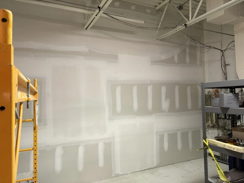 An unfinished interior wall with drywall patches, metal shelving with tools and supplies, a yellow ladder, and exposed ceiling wires.