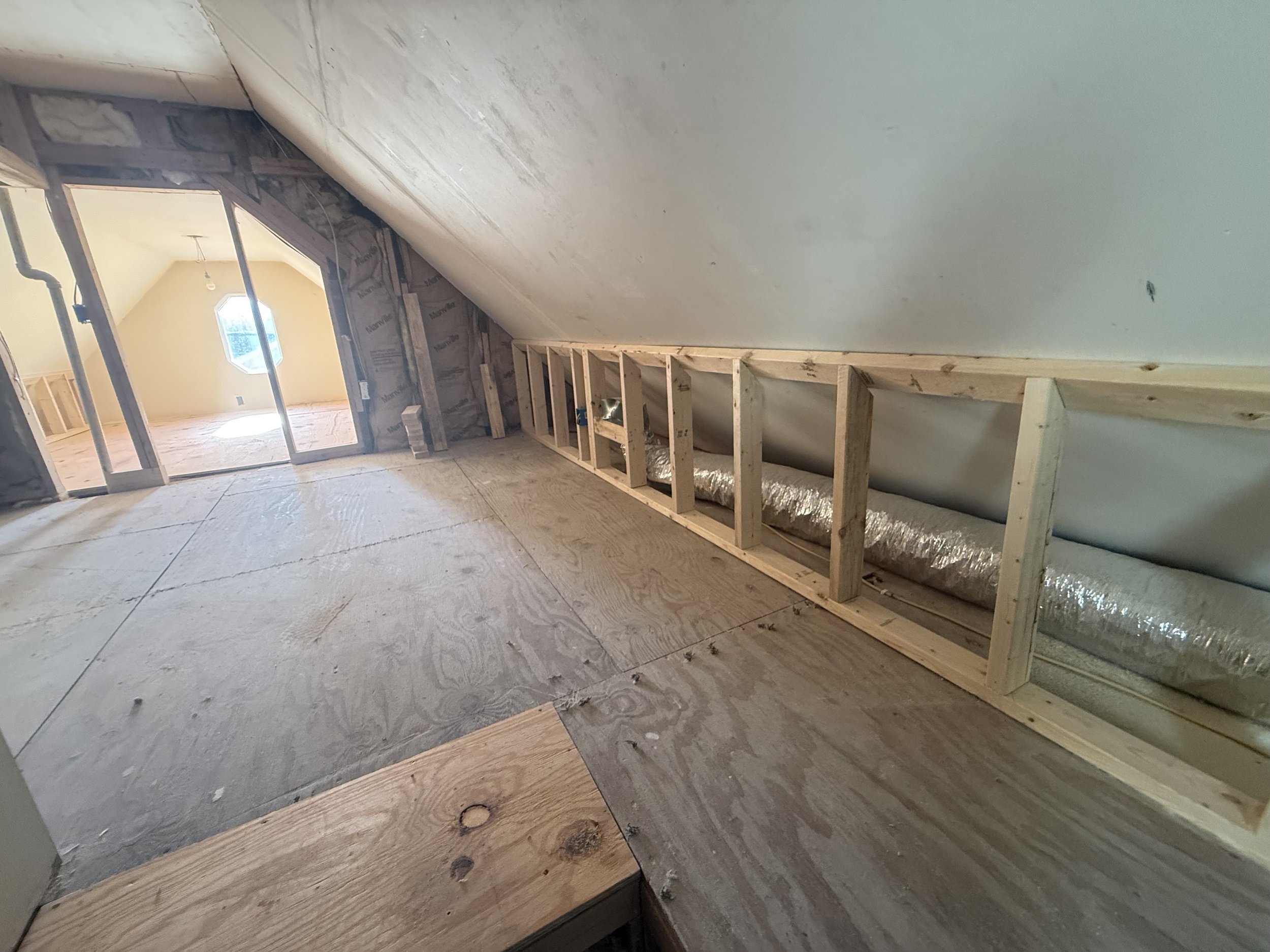 Attic space under construction with exposed drywall, wooden framing, and duct piping, with a small octagonal window in the background.