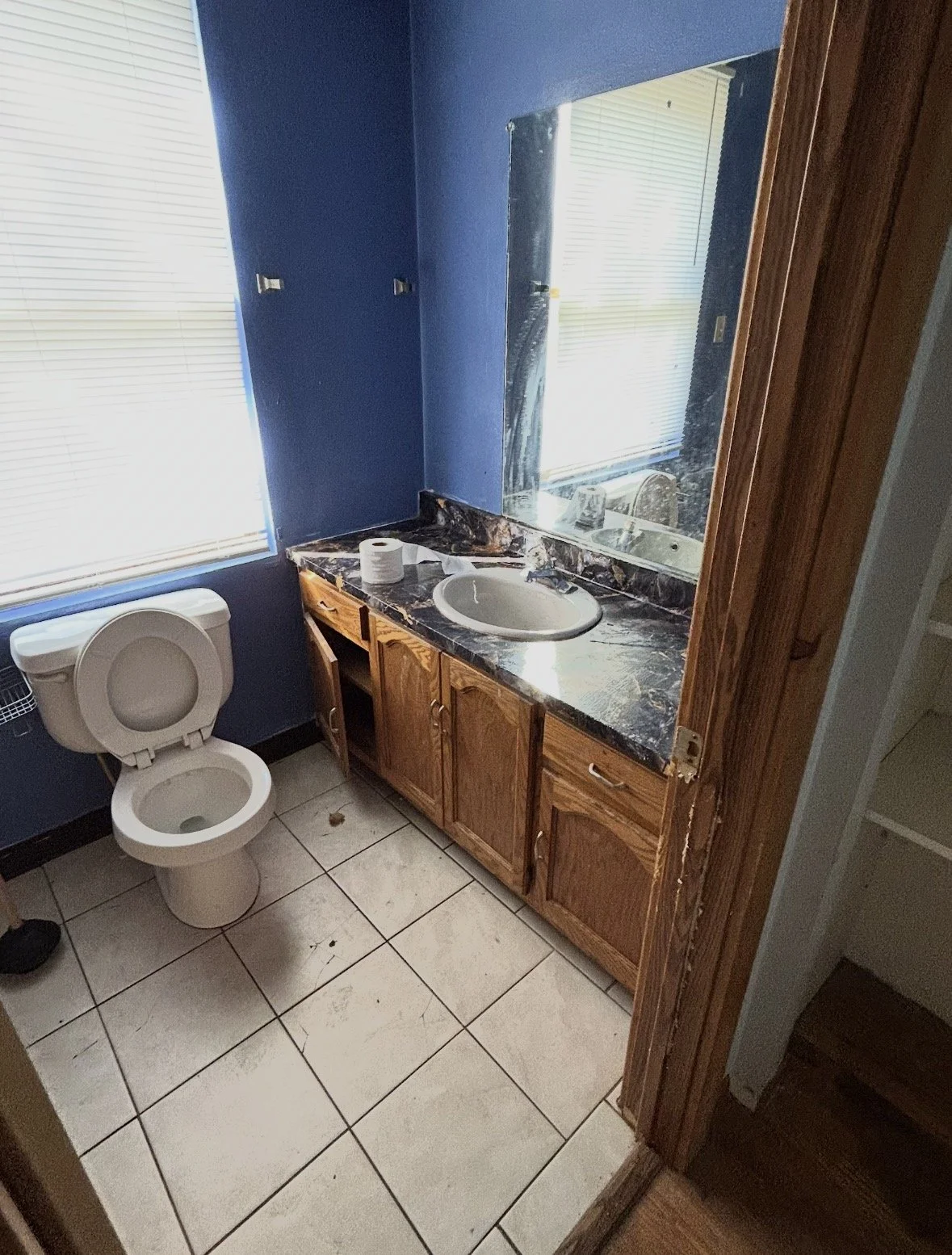 A small bathroom with a blue wall, a window with blinds, a toilet with the lid open, a wooden vanity with a marble countertop, a sink, a mirror, and a roll of toilet paper on the counter.