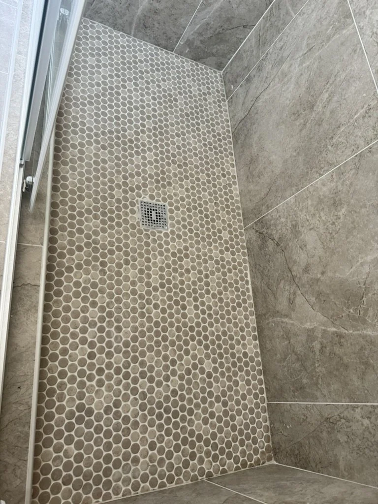 Close-up view of a bathroom shower floor with small, beige hexagonal tiles and a square metal drain in the center, surrounded by large, grayish-brown tiles on the walls.