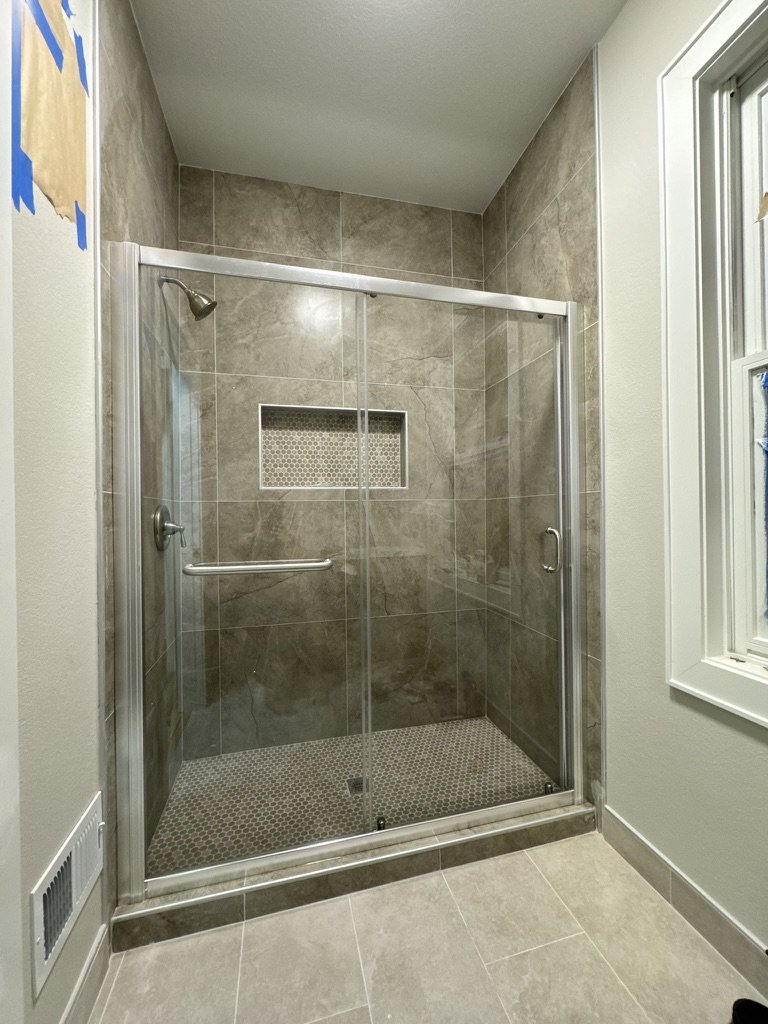 Bathroom shower with glass sliding doors, gray tiled walls, built-in niche, and beige tiled floor.