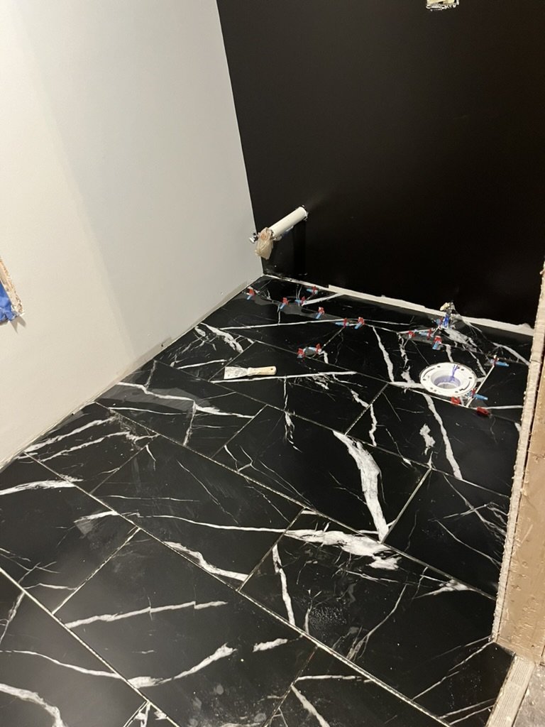Black marble floor with white veining, plumbing pipes without fixtures, and a toilet flange on the floor, indicating ongoing bathroom plumbing work.