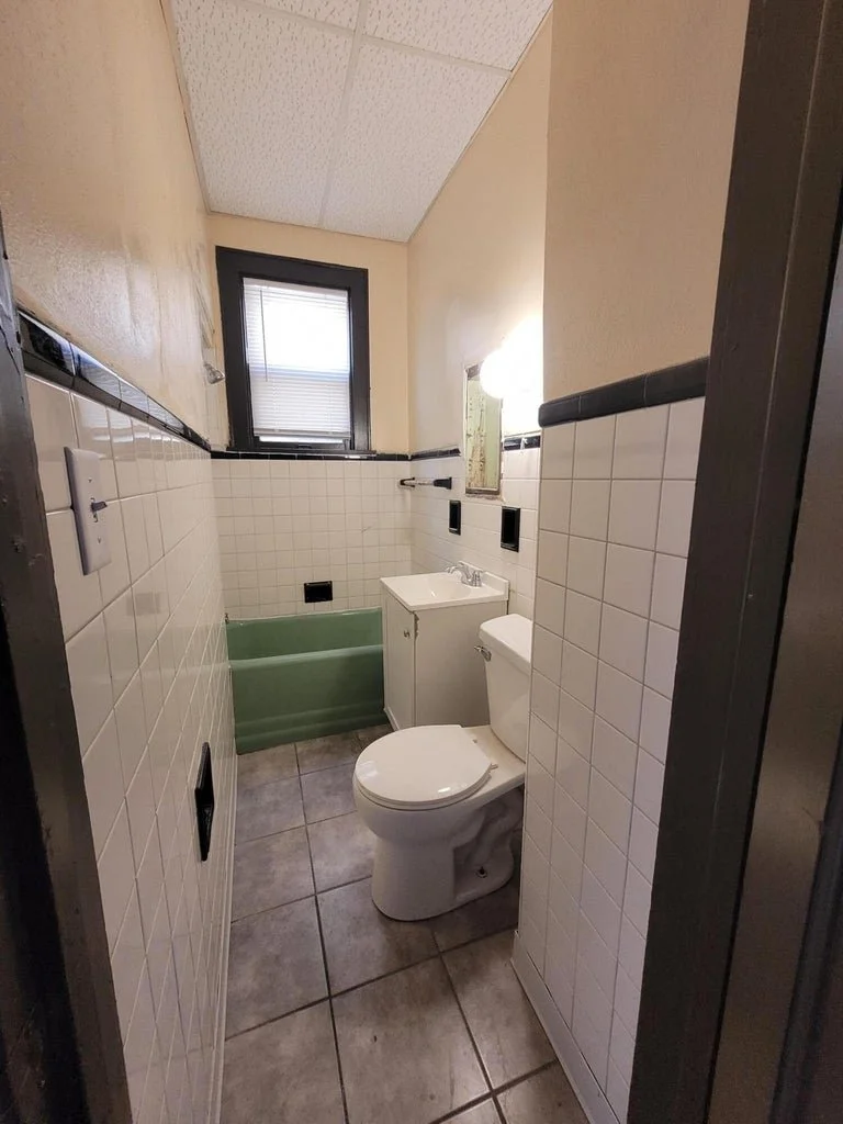 Small bathroom with a toilet, a small sink with cabinet, and a green bathtub. Tiled walls and floor, a window with blinds, and a wall mirror.