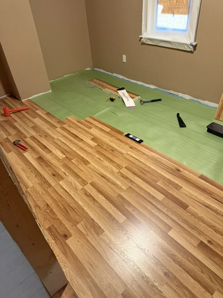 A wooden flooring installation in progress in a room with brown walls and a window. The unfinished floor has some installed hardwood planks, with tools like a hammer, measuring tape, level, and pry bar scattered around. Green paper covers part of the floor, indicating work is ongoing.