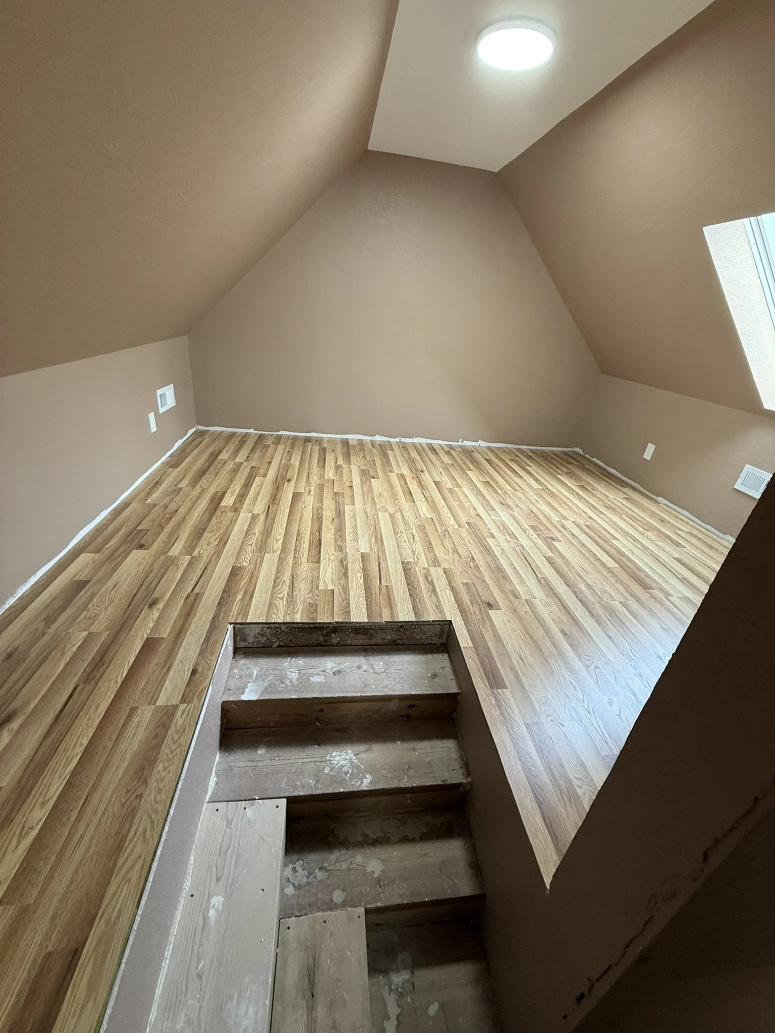 Empty room with sloped ceiling, light brown walls, hardwood flooring, and two skylight windows, with staircase opening visible in the foreground.