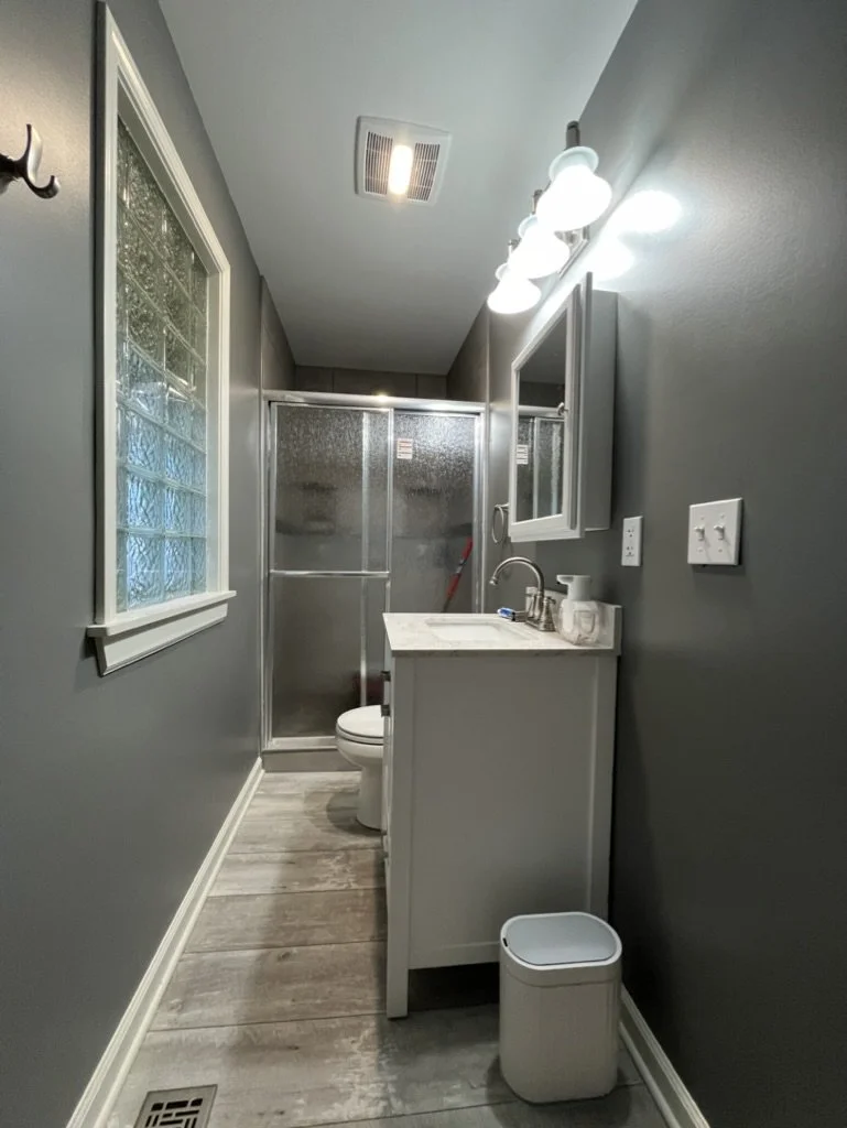 A narrow bathroom with gray walls, a window with glass blocks, a white vanity with a marble top, a mirror cabinet, and a toilet. There is a shower with frosted glass doors and a small trash bin on the floor.