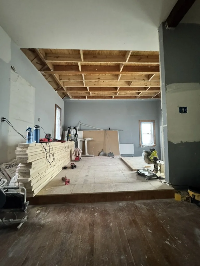 Interior of a room under construction with exposed wooden framing on the ceiling, a small window, and construction tools and materials on a wooden platform.