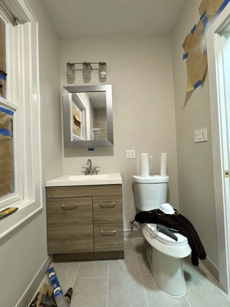 A small bathroom under renovation with a vanity, mirror, toilet, and window. The wall above the toilet has taped paper, and construction tools and paper towels are on the toilet. The floor is tiled, and the walls are painted light gray.