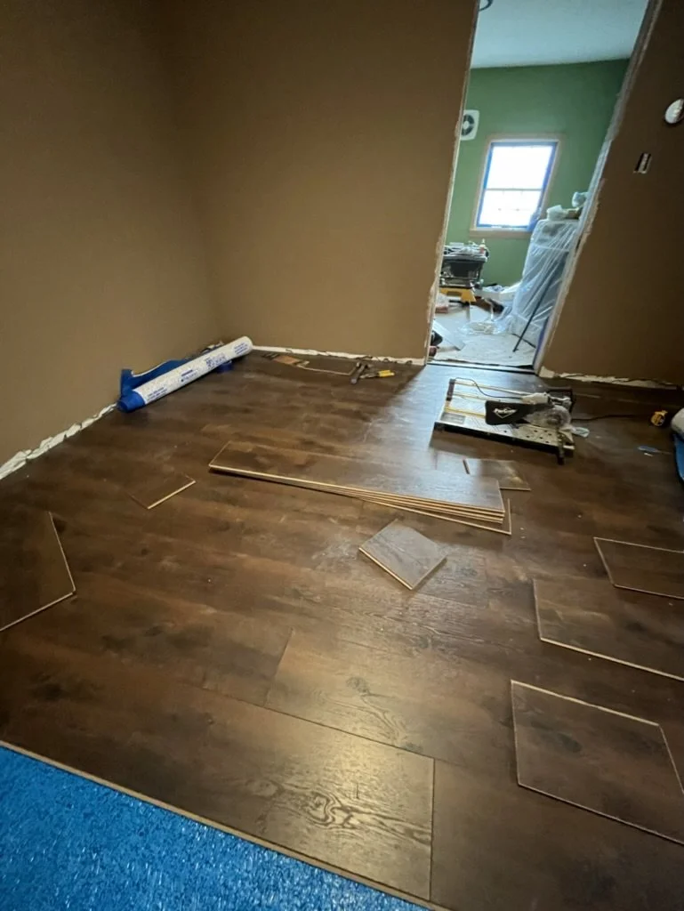 Room under renovation with new hardwood flooring being installed, tiles placed on the floor, drywall and tools visible, doorway leading to a cluttered room with a window, green walls, and construction materials.