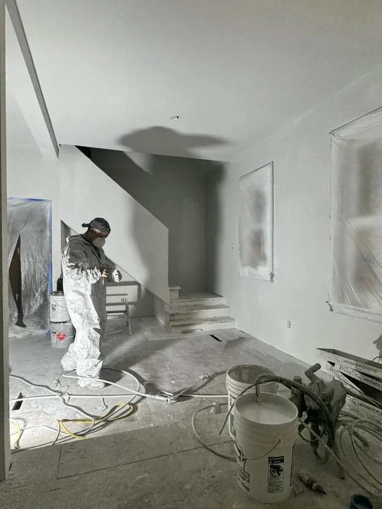 Person in protective clothing working on drywall in a room under construction, with painting supplies, buckets, and electrical cords scattered on the floor.