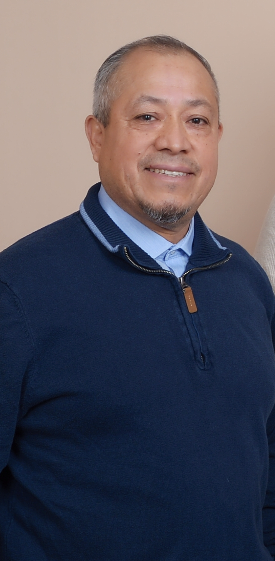 A smiling man with short gray hair, a goatee, wearing a navy blue zip-up sweater over a light blue collared shirt, standing indoors against a beige wall.