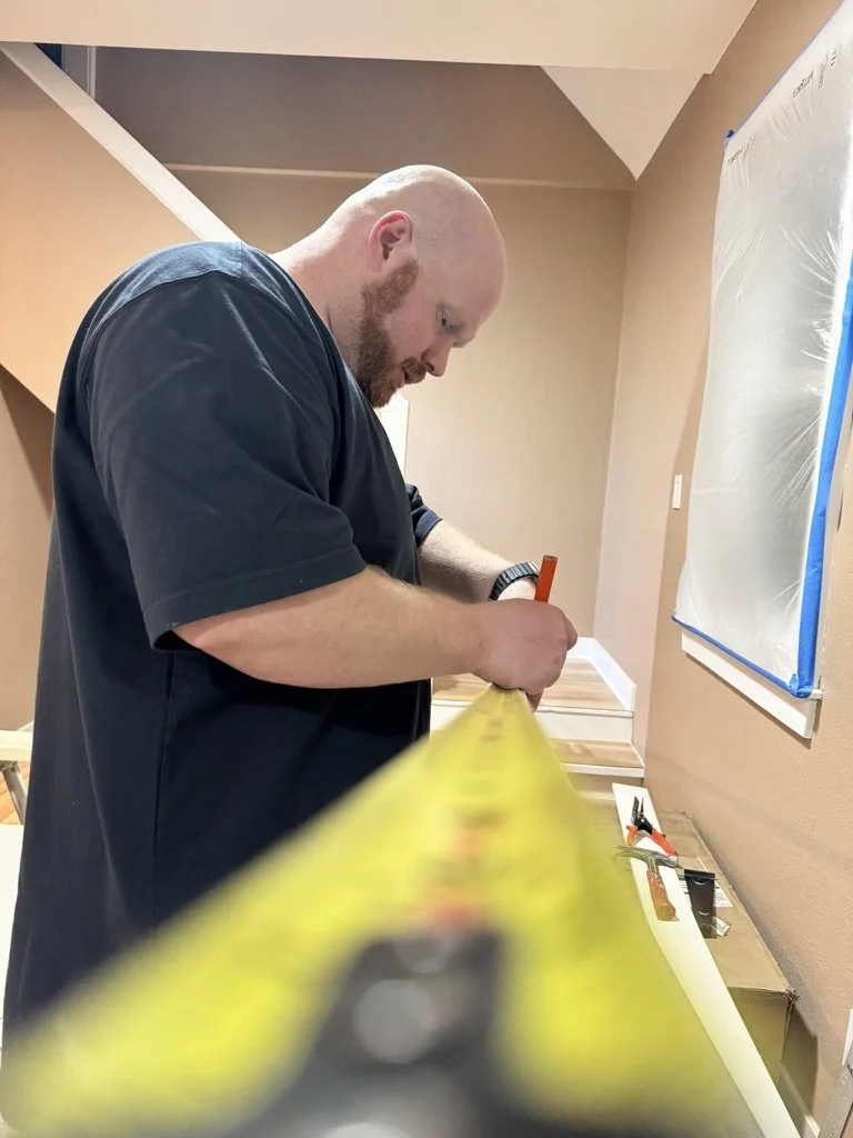 A man with a beard and bald head working on a woodworking project in a room, using a measuring tape and a pencil on a piece of wood, with tools laid out on the table.