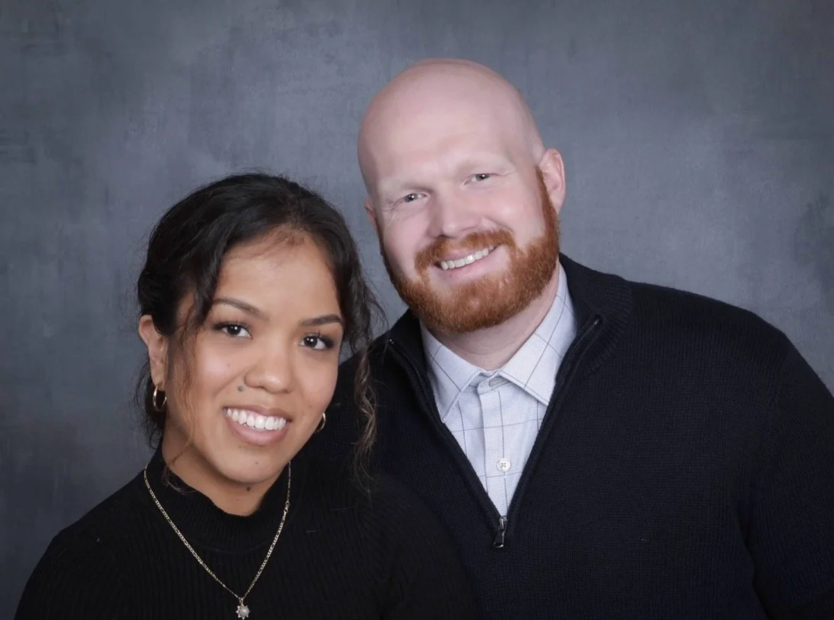 A smiling woman with dark hair and earrings and a man with a beard and bald head, both wearing dark clothing, posing together in front of a gray backdrop.