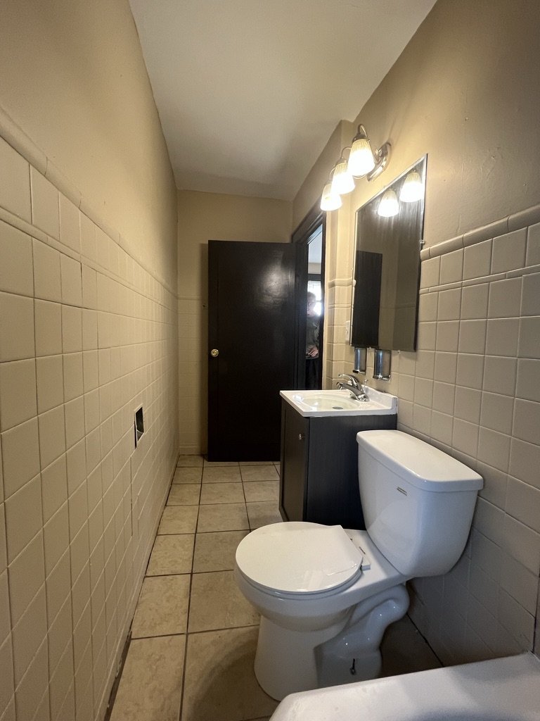Small bathroom with beige tiled walls and floor, a white toilet, a dark cabinet with a sink, and a mirror above with lighting fixtures.