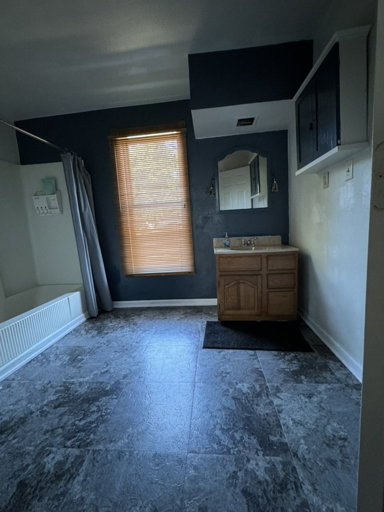 Empty room with a window with blinds, a bathroom sink with a mirror, wall-mounted cabinet, shower area with curtain, and tiled floor.