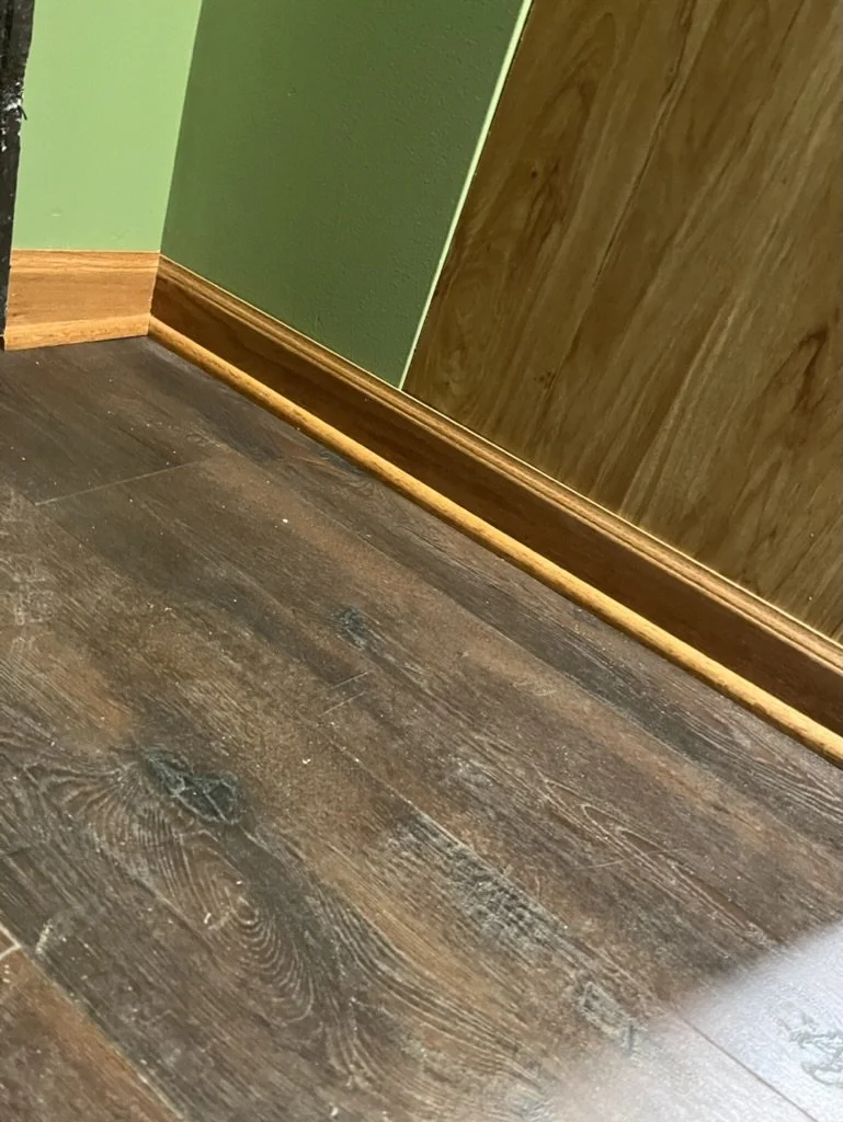 Close-up of a hardwood floor and baseboard against green and wood-paneled walls.
