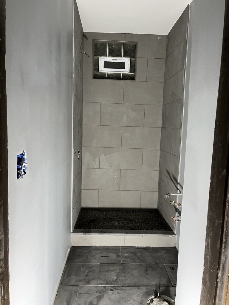 A partially constructed bathroom shower stall with gray tiles on the walls and floor, a small window near the ceiling, and plumbing fixtures for a shower on the right wall.