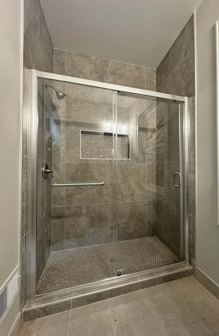 A modern shower with glass sliding doors, gray stone tiles on the walls, small pebble-like tiles on the floor, a built-in niche, and a grab bar.