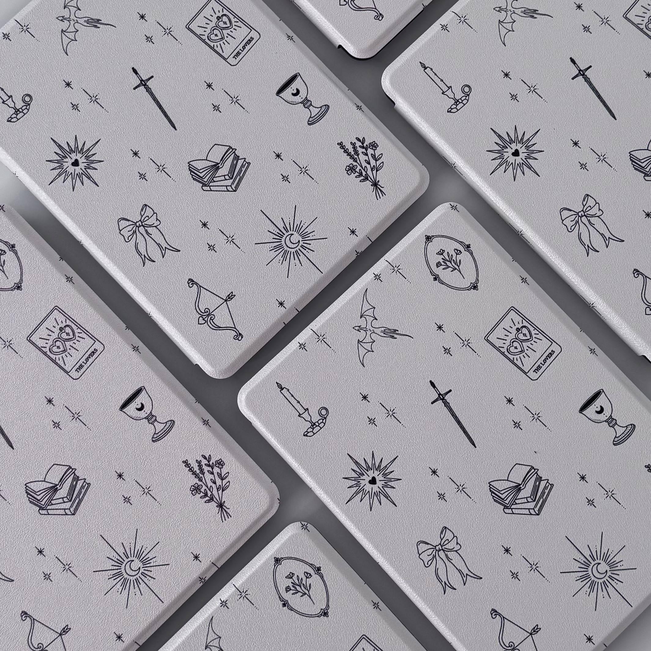 Multiple silver metal cards with black mystical-themed line art illustrations including swords, candles, books, moons, stars, owls, bows, and floral wreaths arranged in a pattern.