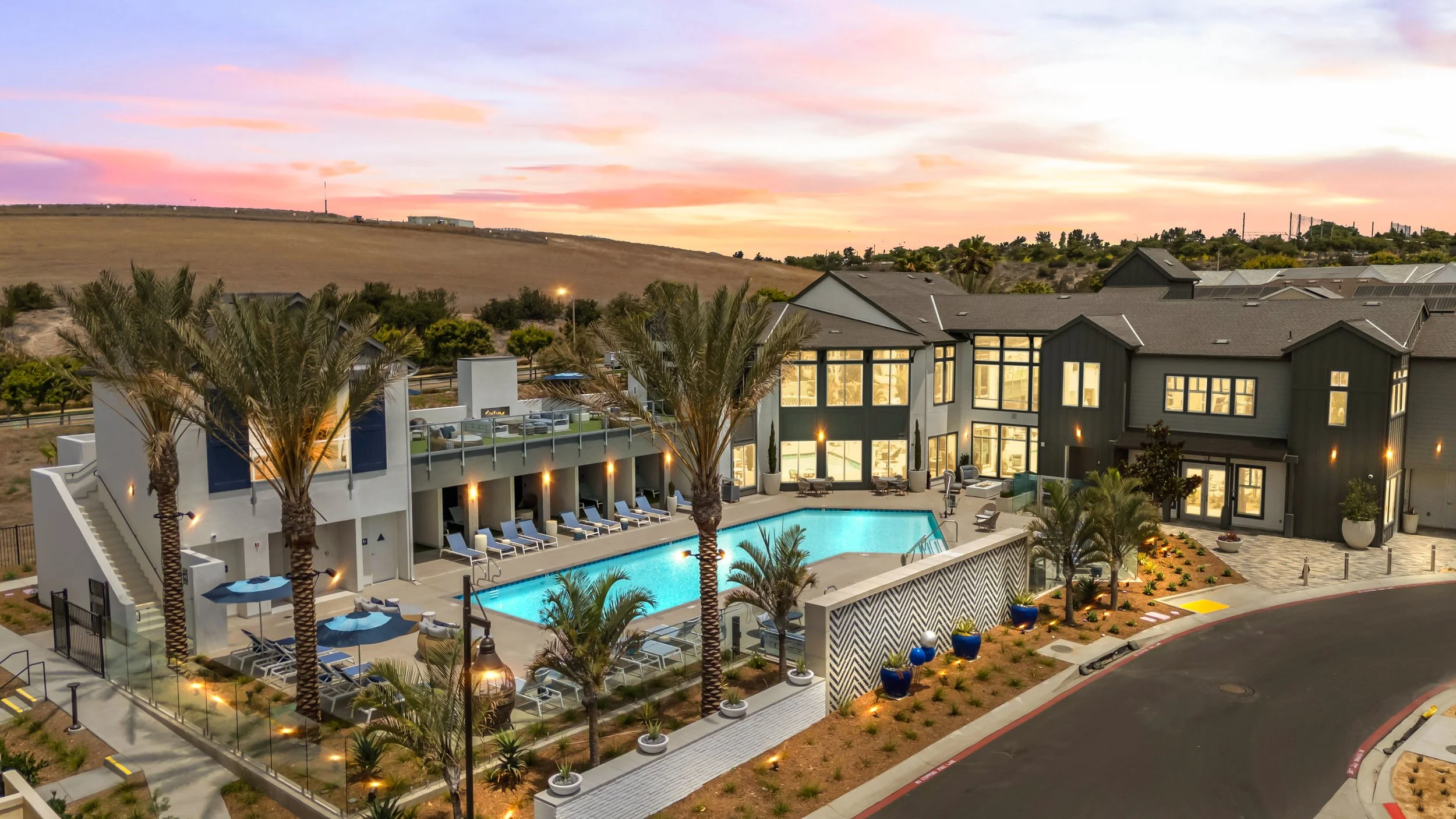 Modern apartment complex with an outdoor swimming pool surrounded by lounge chairs, palm trees, and string lights during sunset.