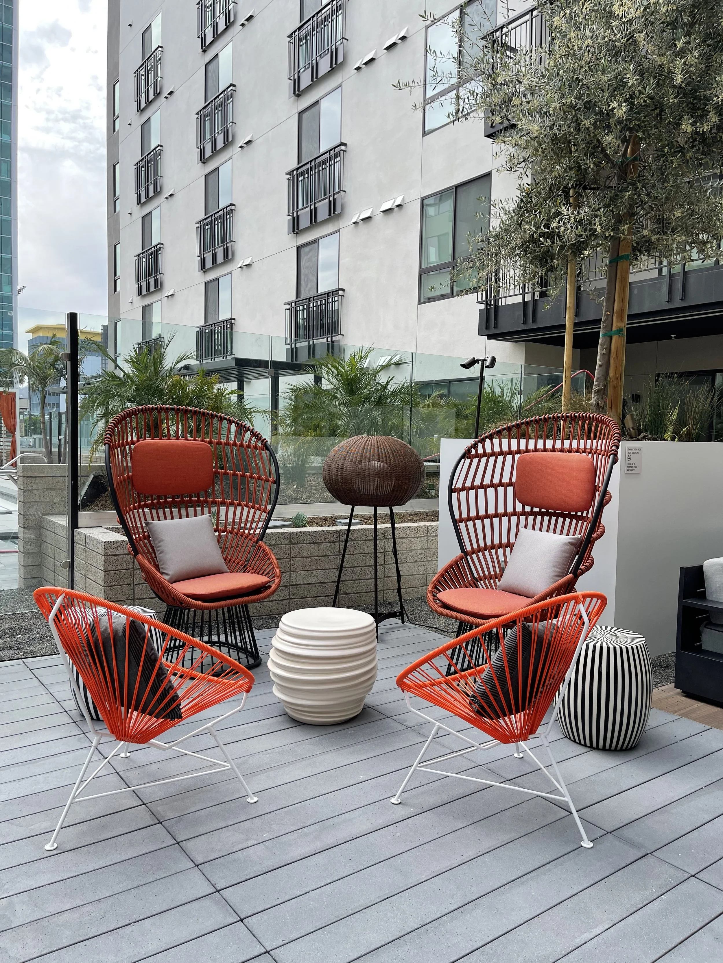 Modern outdoor seating area with four colorful chairs, two are red with black accents, and two are orange with black cushions, around a small round white table, with taller chairs in the background, featuring a cityscape and trees.
