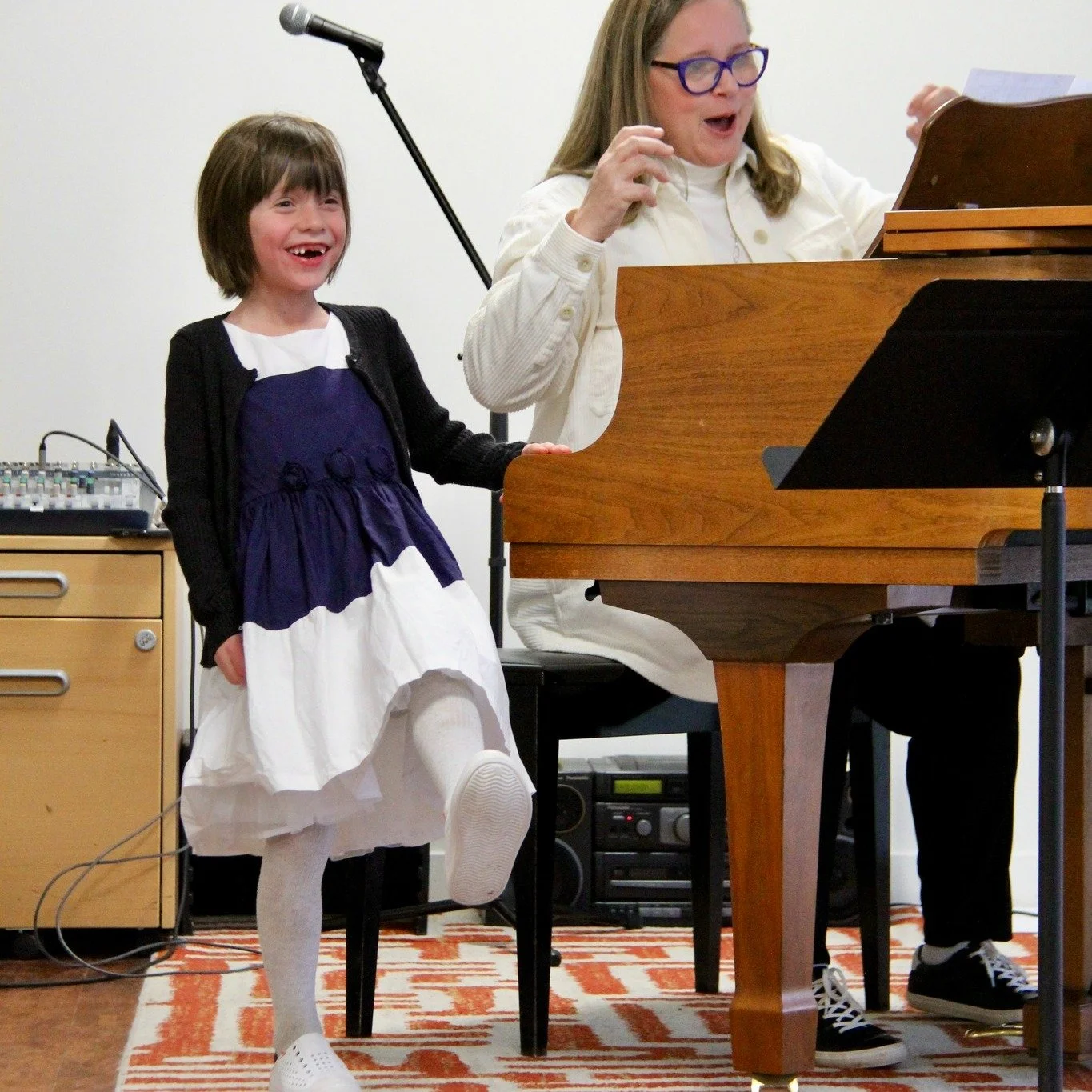 A young girl with short brown hair, wearing a black cardigan, purple and white dress, and white tights, standing next to a woman playing piano. The woman has long hair, glasses, and is dressed in a white shirt. They are performing or practicing music together in a room with a white wall and audio equipment in the background.