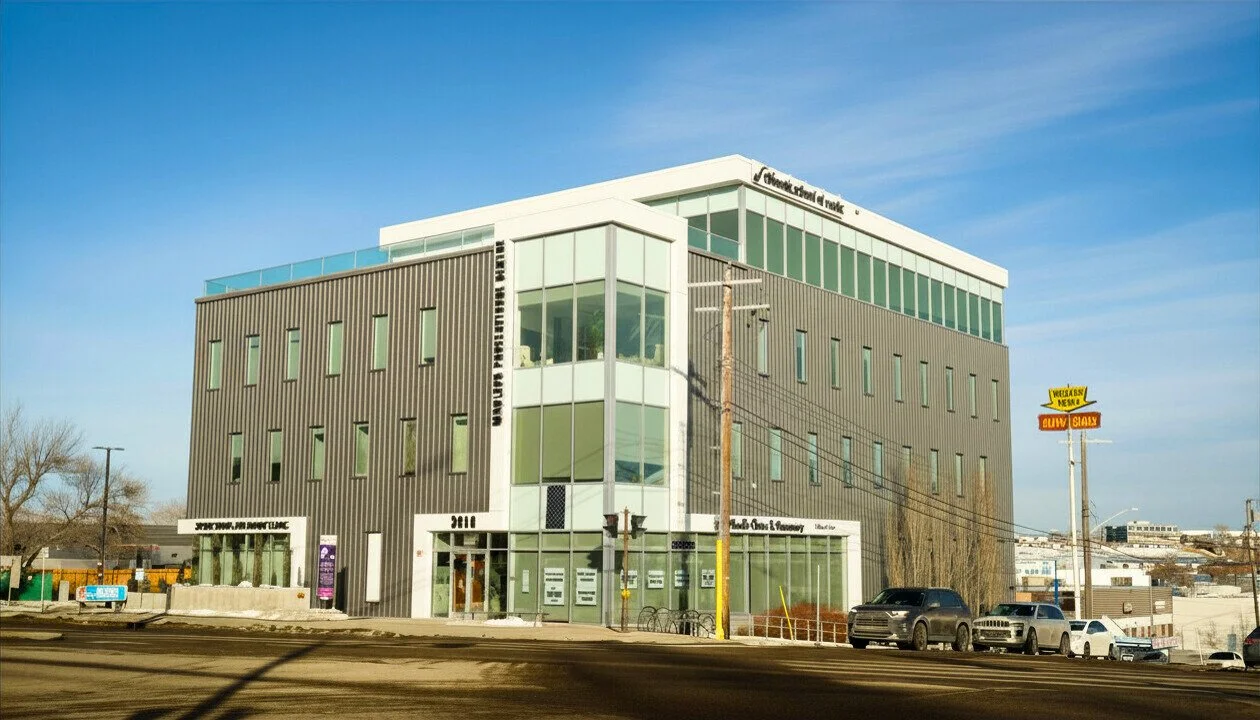 Modern multi-story building with large glass windows and a gray exterior. A street with parked cars and a yellow signboard are in the foreground, with a clear blue sky above.