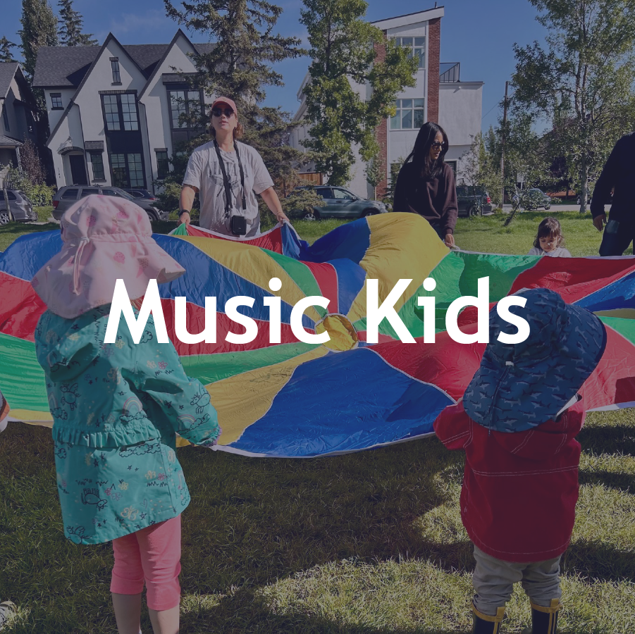 Children and adults playing with a multicolored parachute outdoors on a sunny day. Children are wearing hats and jackets, while adults are helping to lift the parachute. There are residential buildings and trees in the background.
