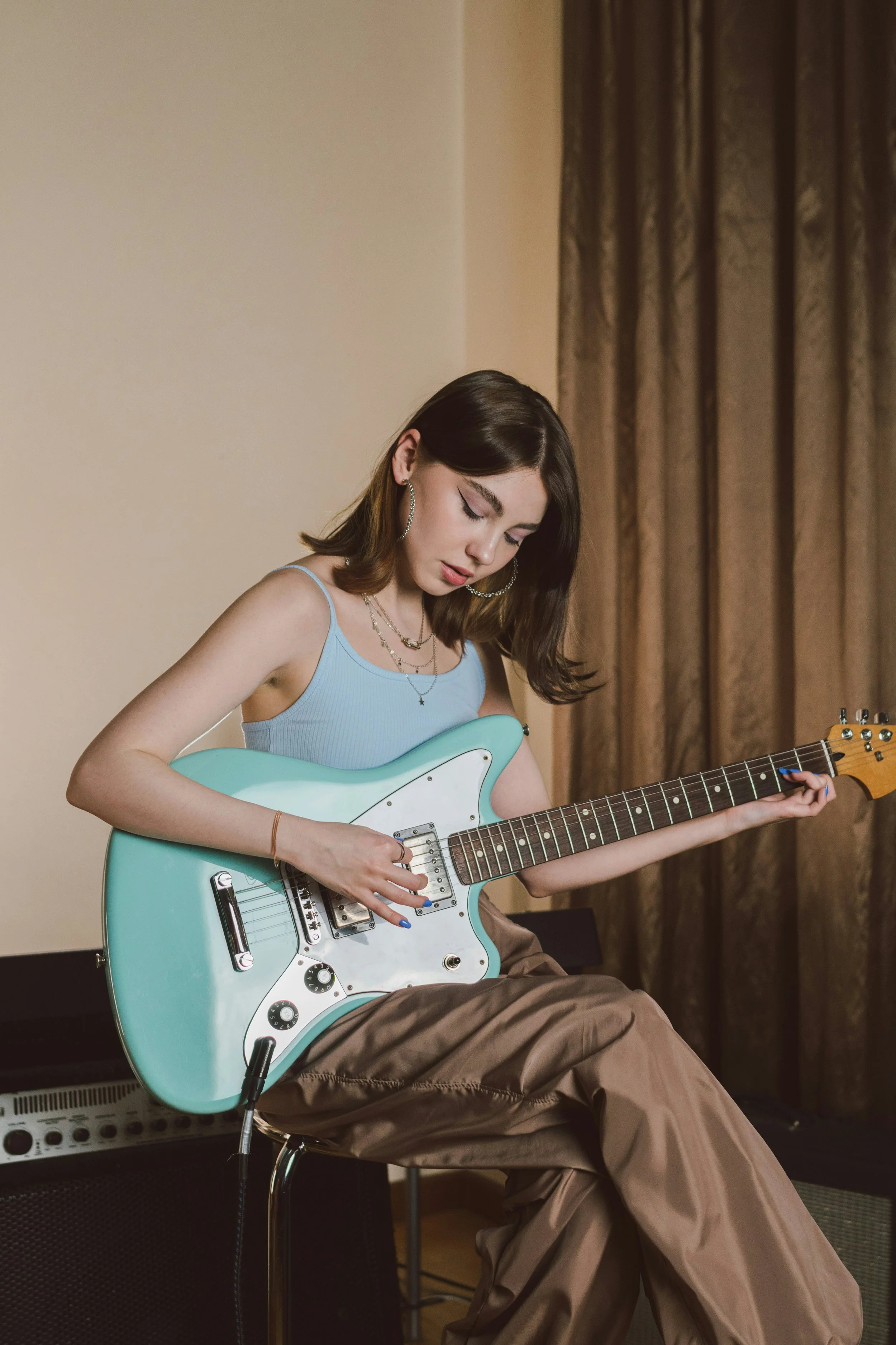 A young woman playing an electric guitar in a room with curtains.