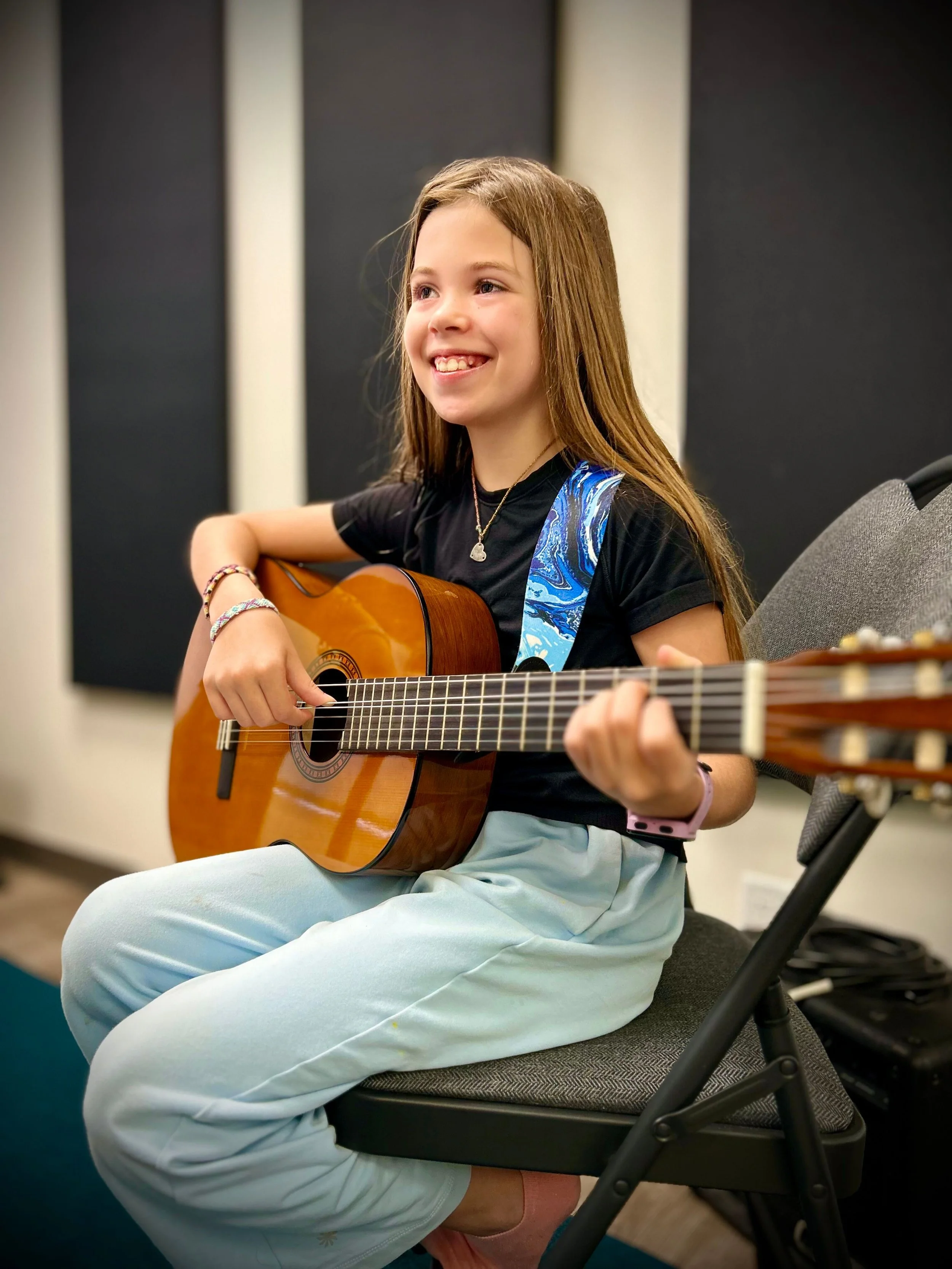 A young girl with long hair playing an acoustic guitar while sitting on a chair in a music room, smiling.