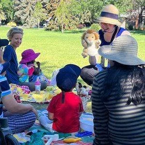 A woman with a hat holding a plush toy and a group of children sitting on a picnic blanket in a park.