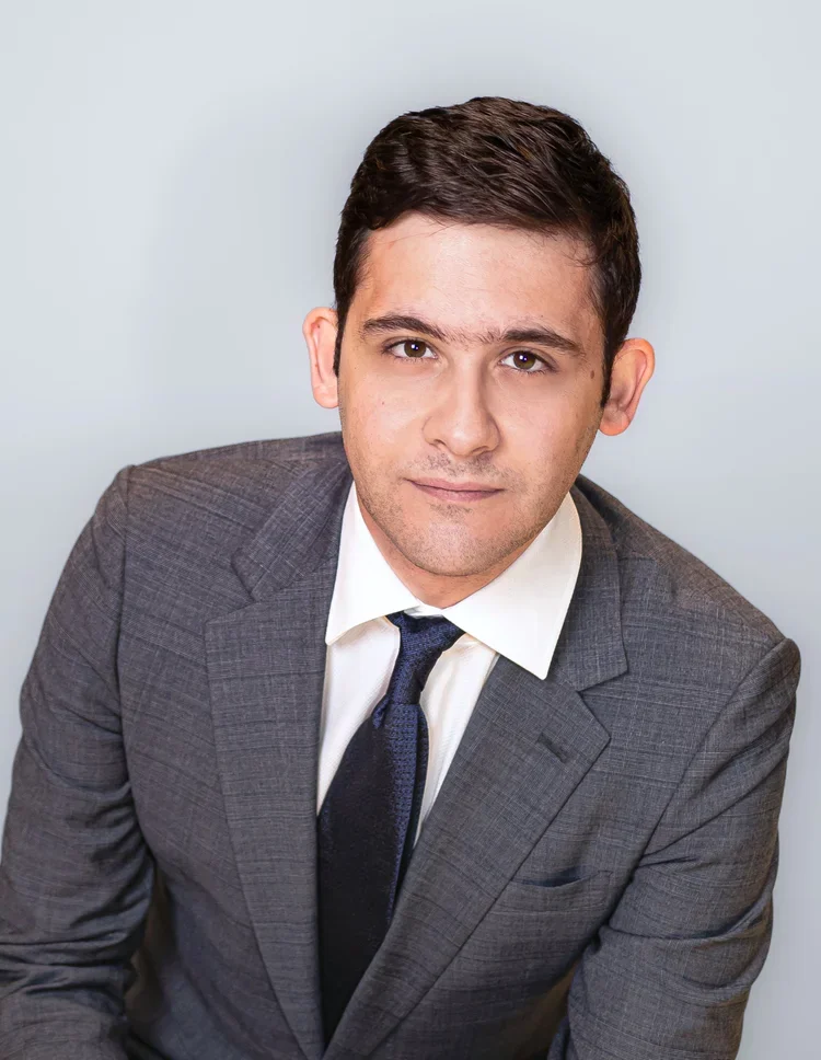 A young man with dark hair, wearing a gray suit, white shirt, and navy tie, posing against a plain light background.
